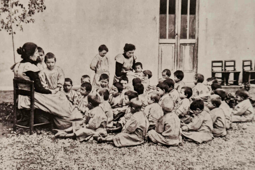 Fotografia del Giardino d'infanzia delle sorelle Agazzi. Le educatrici in giardino attorniate dai bambini.